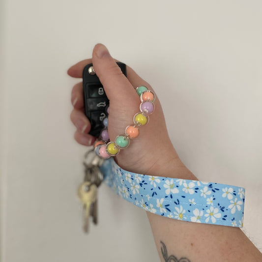 A hand holds a black car key with a pastel beaded wrist strap. The strap is attached to a blue fabric wristlet keyring with a daisy pattern. The tone is casual and playful.