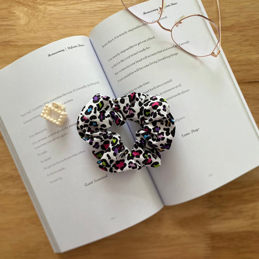 Open book on a wooden table with a colourful leopard-print scrunchie, pearl hair clip, and glasses resting on it, creating a cozy and thoughtful scene.