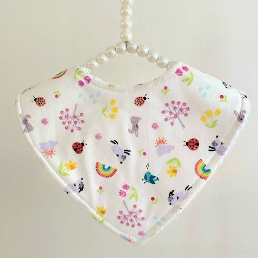 A baby bib with a playful pattern featuring ladybugs, rabbits, sheep, rainbows, and flowers in colourful hues hangs on a beaded hanger against a neutral background.