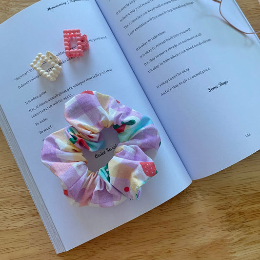 Open book on a wooden table with a pastel and strawberry print scrunchie and beaded hair clips on the pages, next to gold wire glasses, creating a cozy, creative vibe.